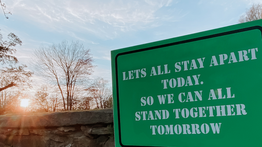 Green sign in front of a manmade rock wall that says Let's All Stay Apart Today. So We Can All Stand Together Tomorrow